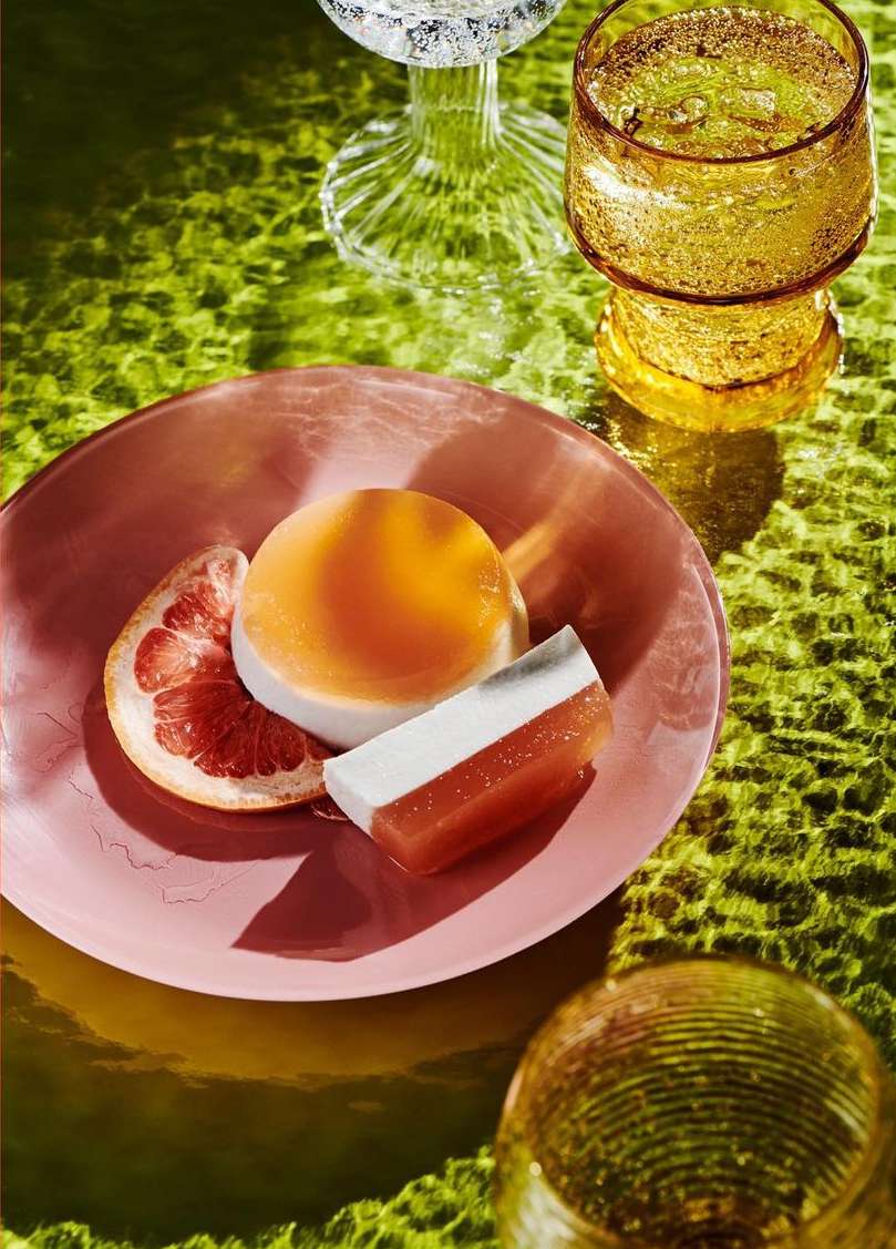 Studio shot of pink plate with citrus fruit slice and layered dessert, surrounded by sparkling drinks on a patterned surface.