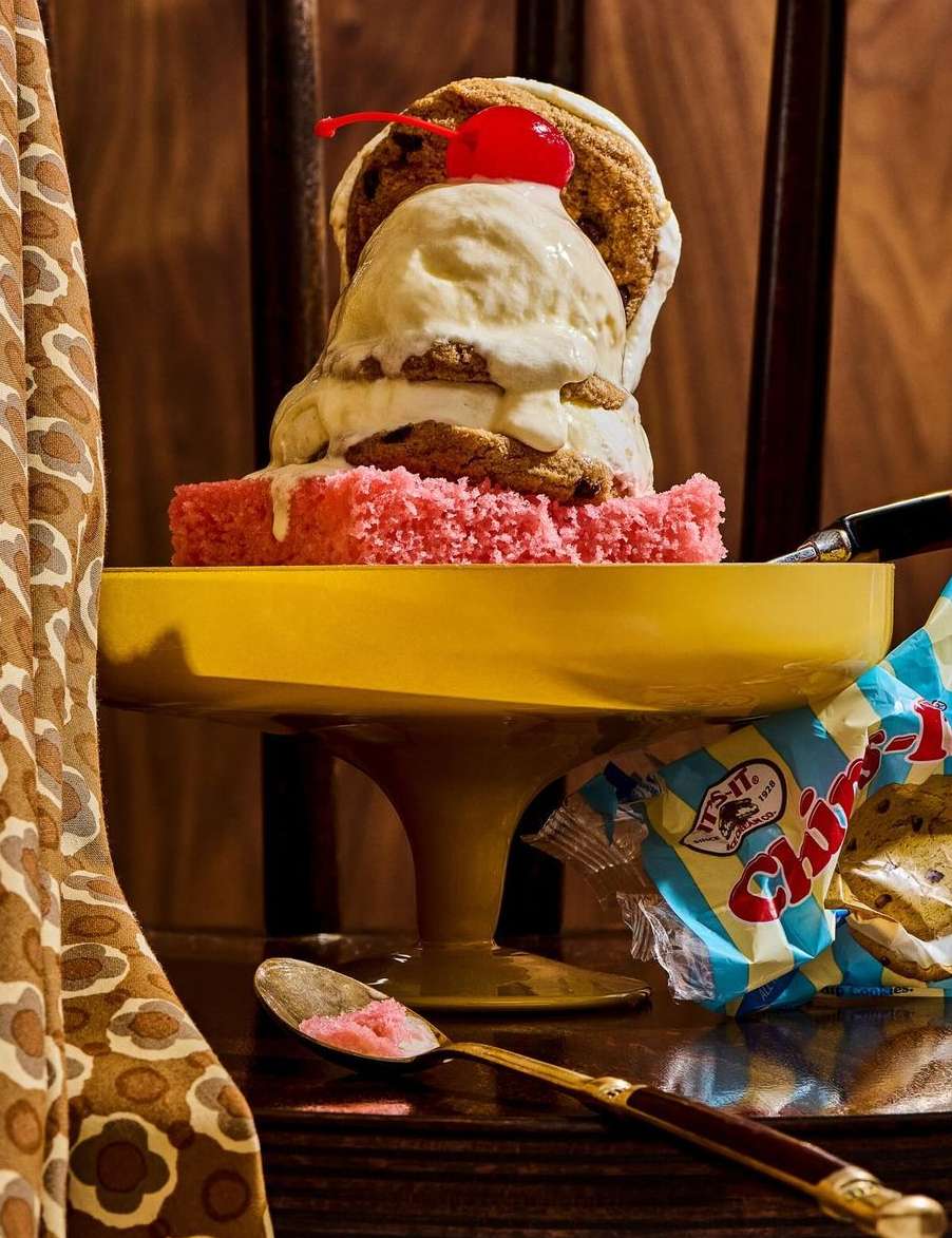 A dessert featuring a cake topped with scoops of ice cream and a cherry, presented on a yellow cake stand against a wooden background.