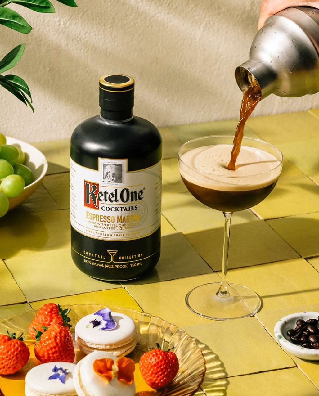 Pouring espresso martini cocktail from shaker into glass next to liquor bottle, macarons, strawberries, grapes, and coffee beans.