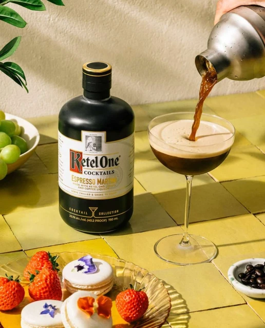 Pouring espresso martini cocktail from shaker into glass next to liquor bottle, macarons, strawberries, grapes, and coffee beans.