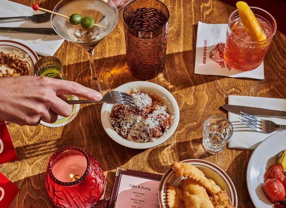 Overhead shot of a wooden table featuring cocktails, a candle, bowls of food, and a person holding a fork, conveying a cozy atmosphere.