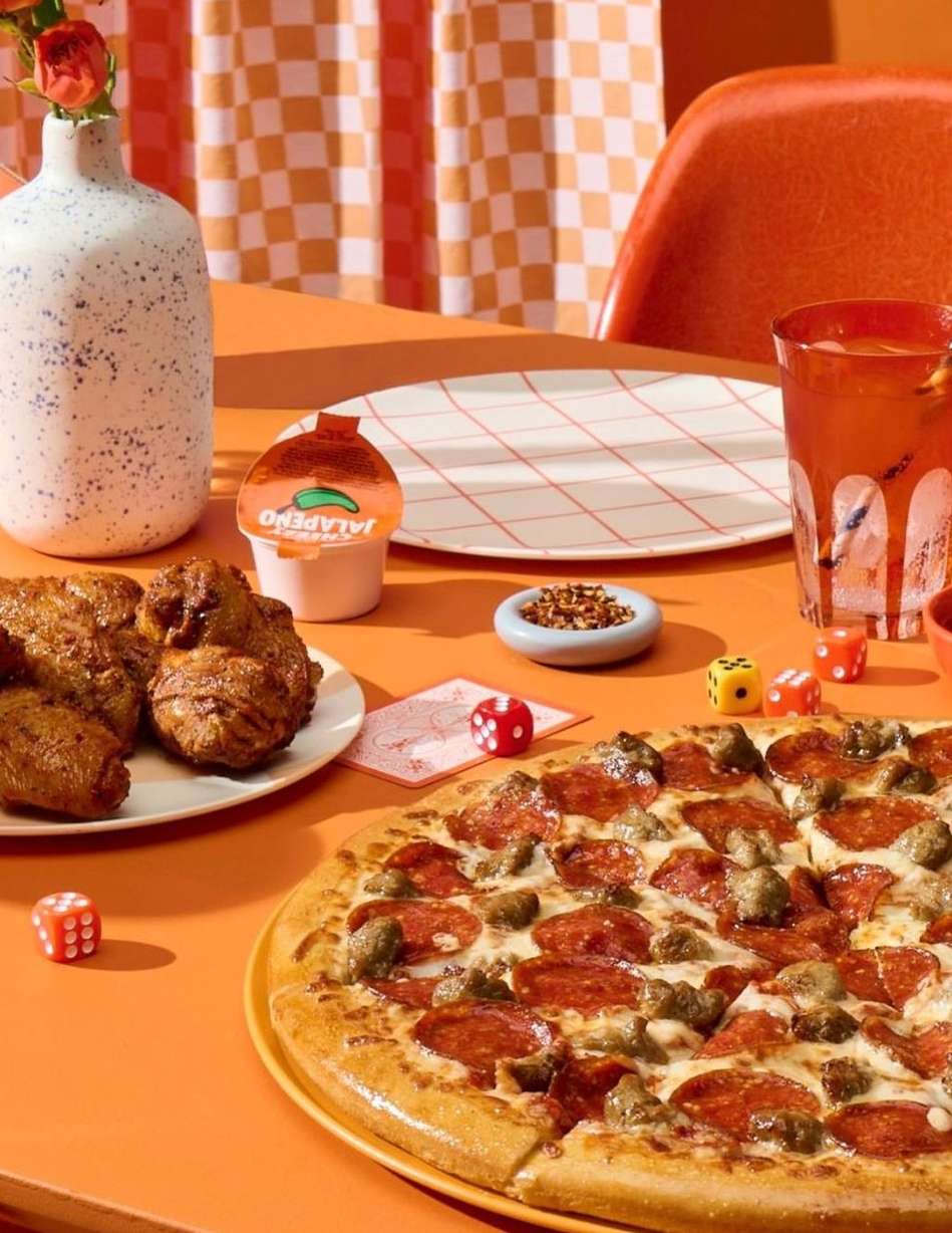 Overhead shot of pepperoni pizza, fried chicken, table with orange and white checkered tablecloth, drink, vase with flowers, and dice.