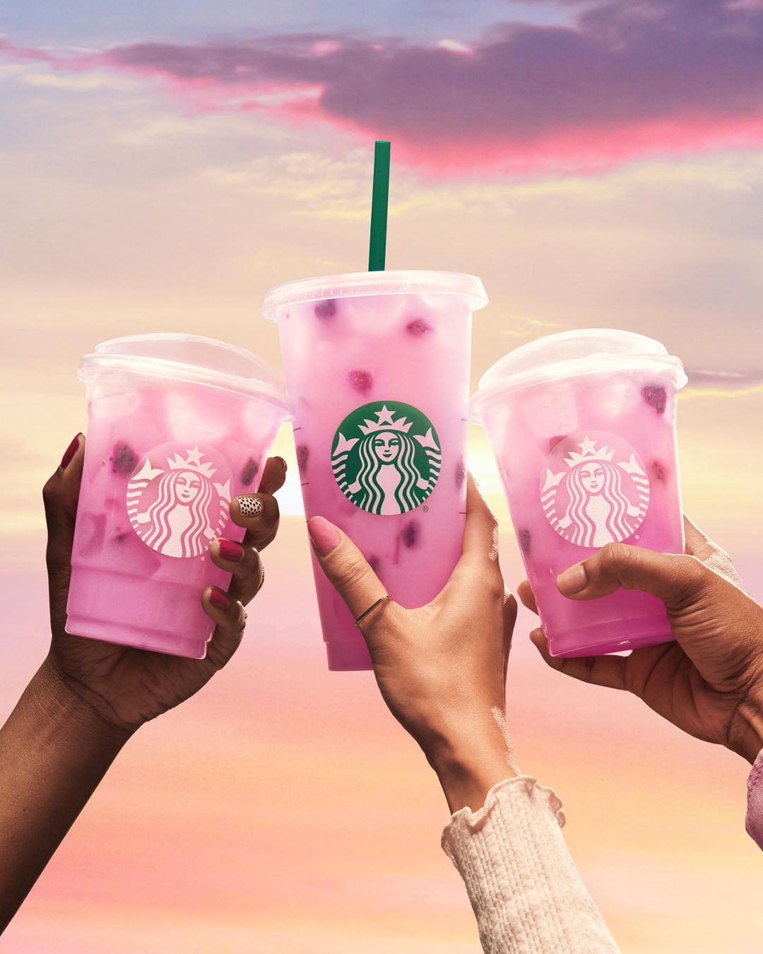 Three Starbucks cups with pink drinks are held up by arms against a colorful sky, perfect for social media marketing in food and beverage.