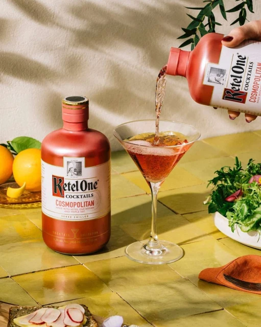 Pouring Ketel One Cosmopolitan cocktail into martini glass, featuring vibrant colors, citrus fruits, and culinary presentation.