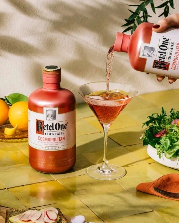 Pouring Ketel One Cosmopolitan cocktail into martini glass, featuring vibrant colors, citrus fruits, and culinary presentation.
