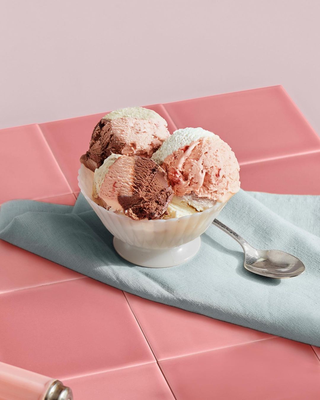 Close-up of a bowl of three scoops of chocolate, vanilla, and strawberry ice cream served on a pink tile background with a spoon.