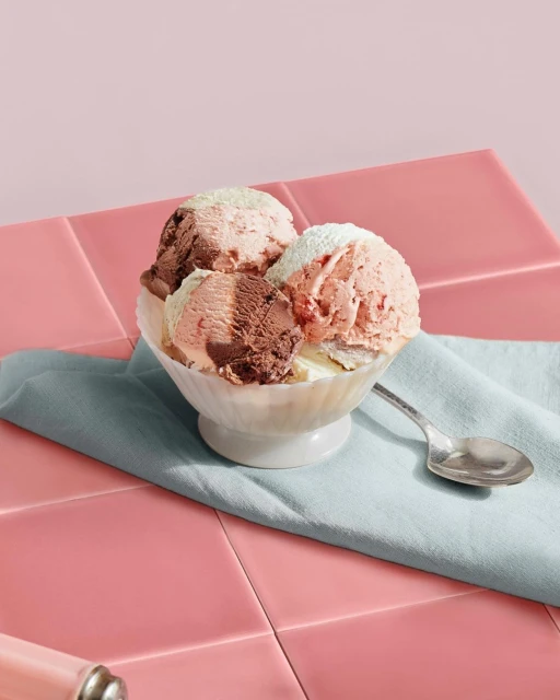 Close-up of a bowl of three scoops of chocolate, vanilla, and strawberry ice cream served on a pink tile background with a spoon.
