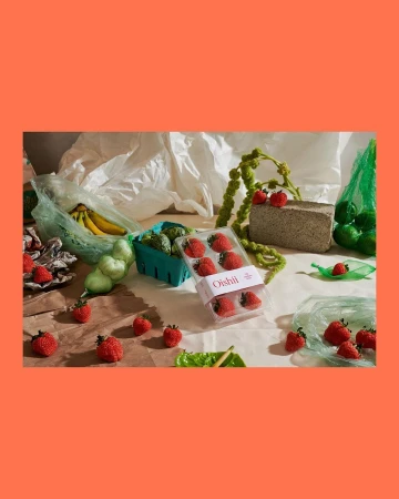 Still life of strawberries in packaging, bananas, limes, and other groceries arranged on a neutral surface against a coral background.