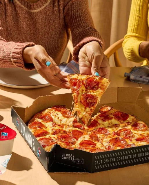 Close-up of a pepperoni pizza in a Domino's box, an adult hand is lifting a slice with cheese dripping from the crust at the table.