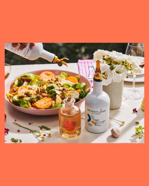 Pouring olive oil onto a salad with Brightland olive oil bottles, flowers, and glasses on a white table with an orange background.