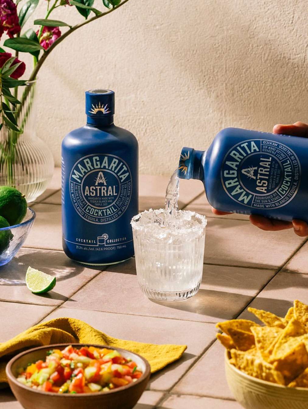 Margarita Astral cocktail being poured into a salt-rimmed glass, surrounded by limes, tortilla chips, and salsa on a tiled surface.