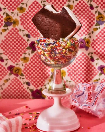 Ice cream sandwich on a glass full of colorful sprinkles, resting on a pink and white stand, against a patterned background.