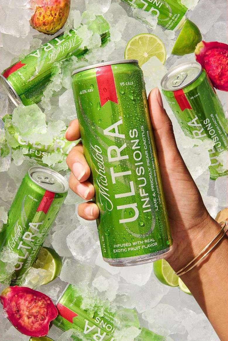 Michelob Ultra Infusions can held by a person's hand, surrounded by ice cubes, lime wedges, and pieces of dragon fruit.