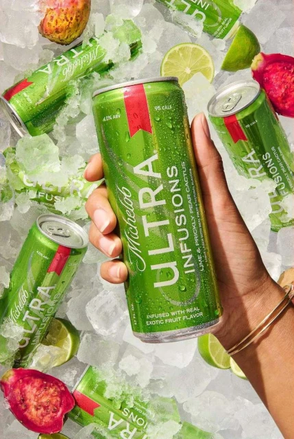 Michelob Ultra Infusions can held by a person's hand, surrounded by ice cubes, lime wedges, and pieces of dragon fruit.
