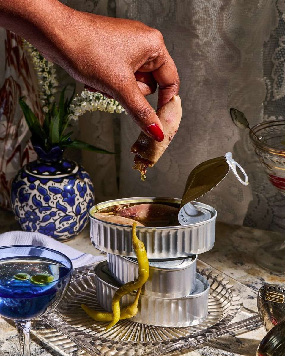 Hand holding stuffed pastry over stacked sardine cans, glass of blue liquid, patterned tablecloth, and vase with white flowers.