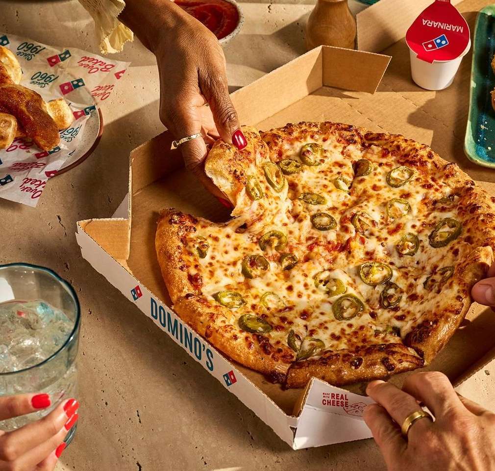 Overhead shot of a Domino's pizza with jalapeños being served. Several hands reach to take a slice, surrounded by other food items.