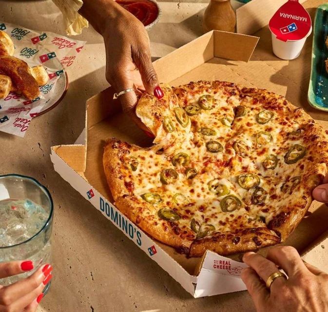 Overhead shot of a Domino's pizza with jalapeños being served. Several hands reach to take a slice, surrounded by other food items.