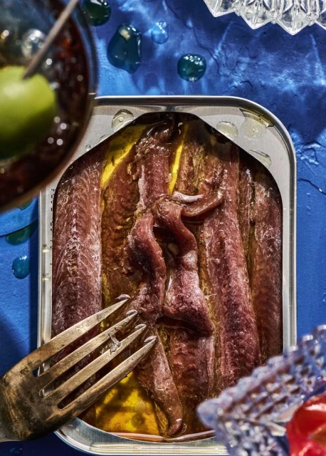 Overhead shot of a silver can of anchovies in oil, garnished with a fork, olive, and crystal glass on textured blue background.