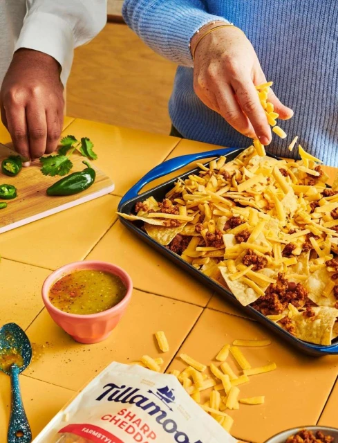 Close-up of hands preparing nachos with tortilla chips, ground meat, shredded cheese, salsa, cilantro, and jalapeños on a yellow tile surface.