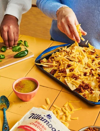Close-up of hands preparing nachos with tortilla chips, ground meat, shredded cheese, salsa, cilantro, and jalapeños on a yellow tile surface.