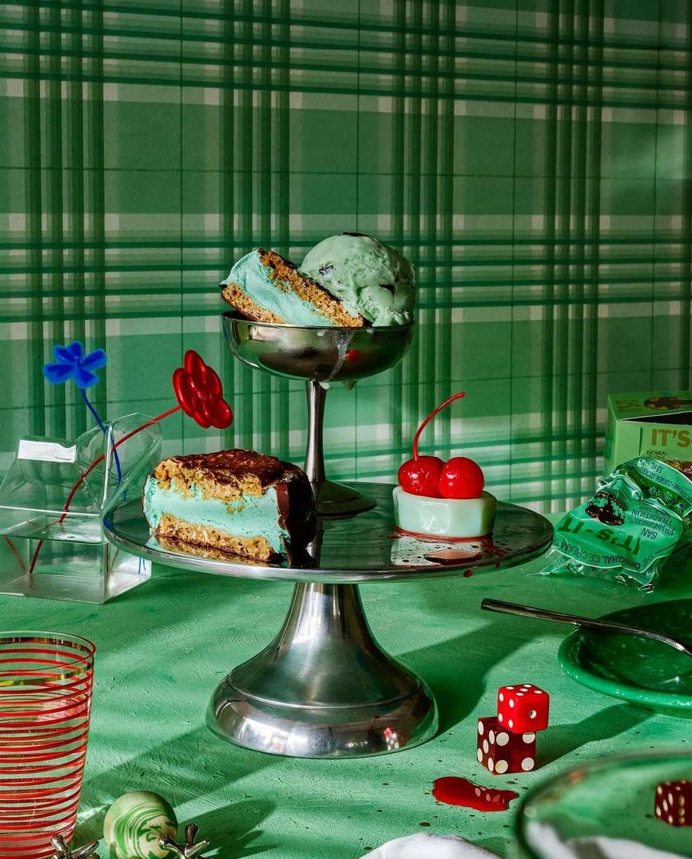 Still life with ice cream sandwiches, mint chip ice cream, cake, and dice on a silver cake stand with a green patterned wallpaper.