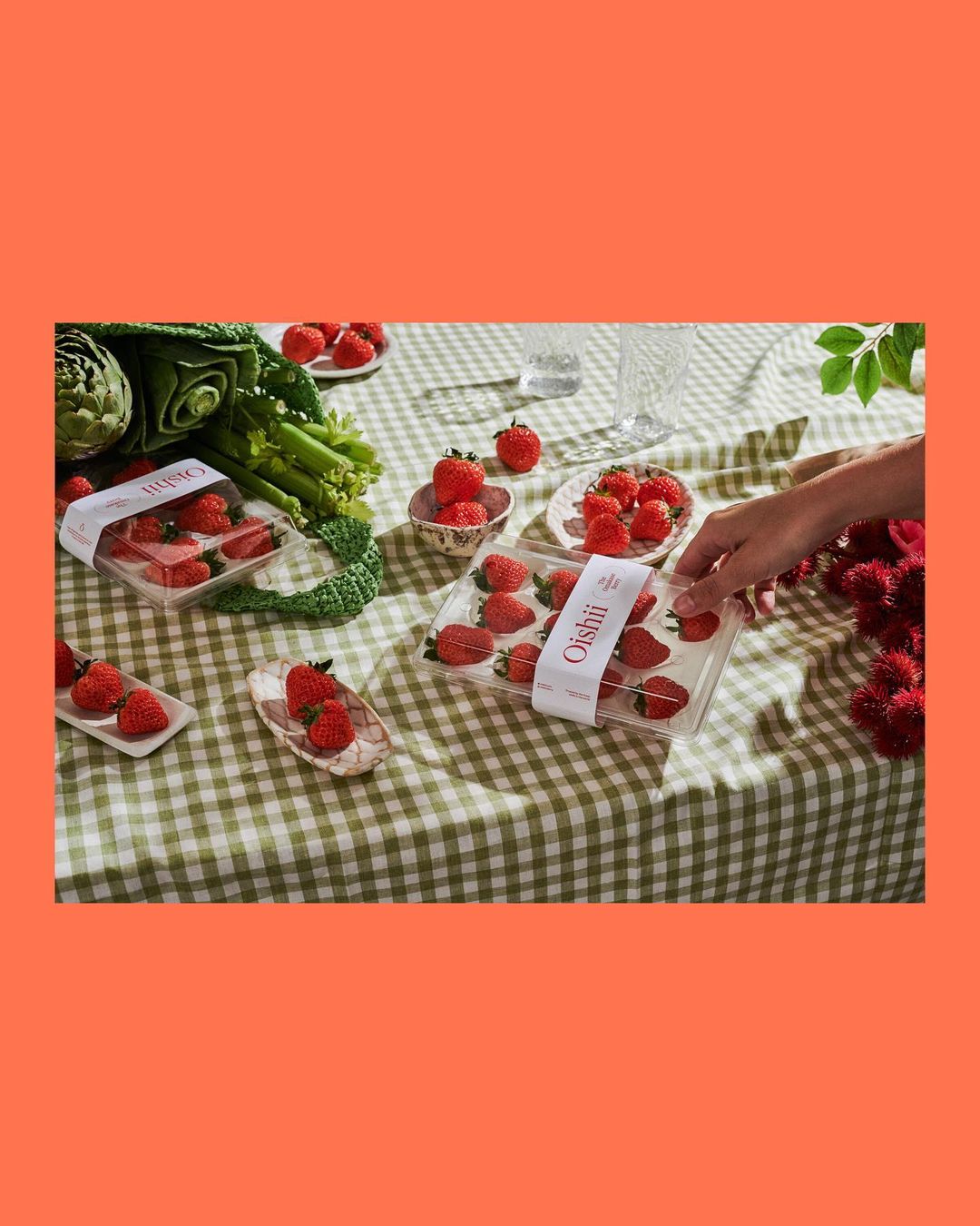Overhead shot of fresh strawberries in plastic containers on a green checkered tablecloth, with vegetables, a hand, and glasses.