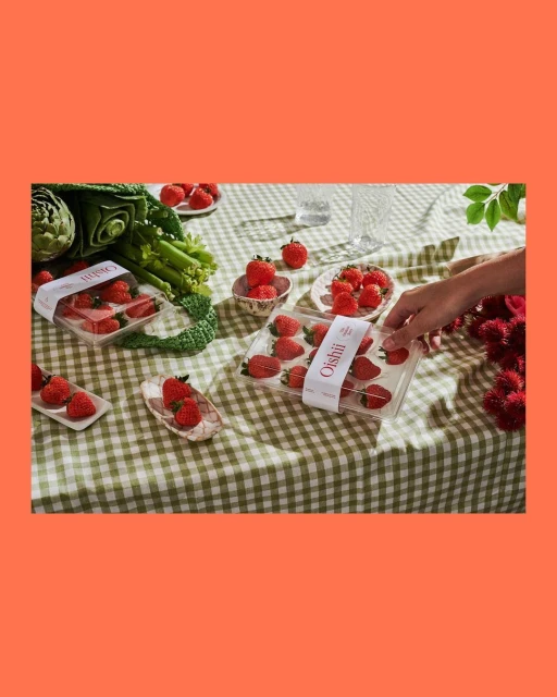 Overhead shot of fresh strawberries in plastic containers on a green checkered tablecloth, with vegetables, a hand, and glasses.