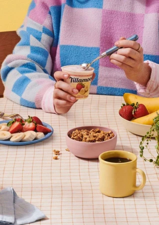 Person eating yogurt at a table with fruit and a beverage, wearing a colorful sweater, and holding a spoon in a cozy indoor setting.
