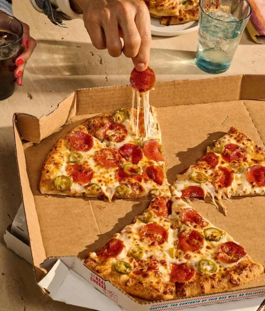 Overhead shot of pepperoni pizza slices in a cardboard box with a hand holding a pepperoni slice with stretchy cheese.