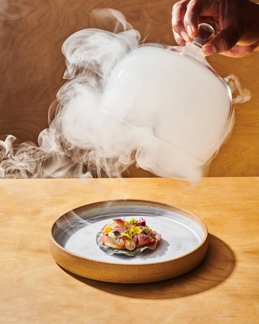 Overhead shot of a chef serving a dish on a ceramic plate with a smoke-filled glass cloche, set on a wooden table in a studio environment.