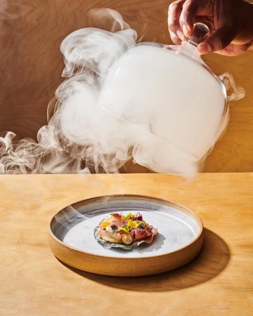 Overhead shot of a chef serving a dish on a ceramic plate with a smoke-filled glass cloche, set on a wooden table in a studio environment.