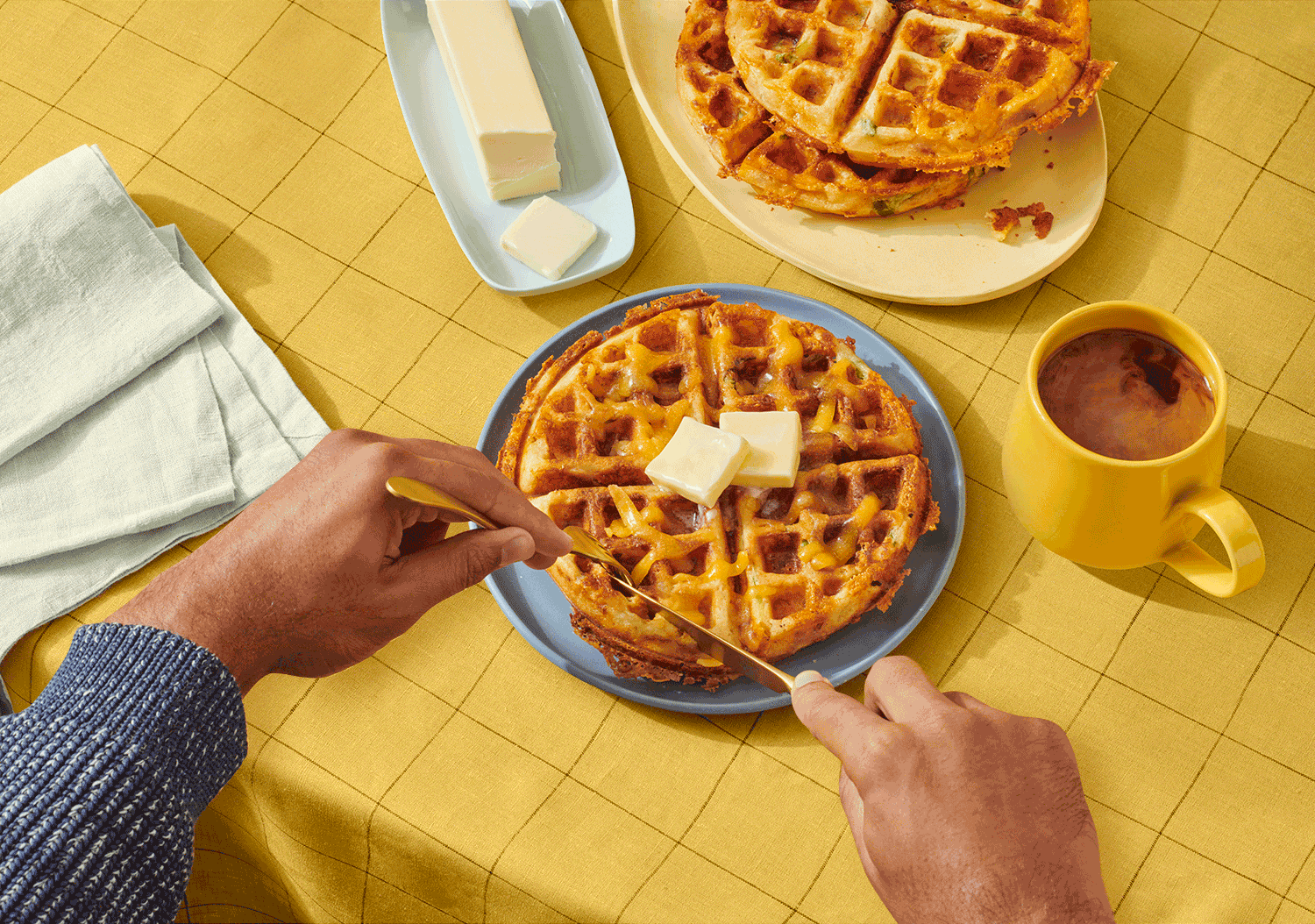 Overhead shot of a person cutting into a golden waffle topped with butter at a table with a yellow tablecloth and light blue plates.