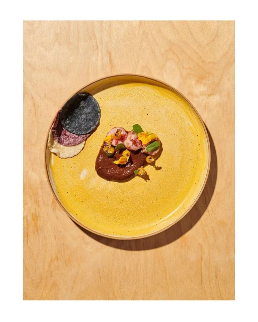 Overhead shot of an artfully plated appetizer on a yellow plate, featuring a dark sauce and colourful toppings, set against a wood background.