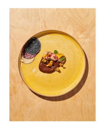 Overhead shot of an artfully plated appetizer on a yellow plate, featuring a dark sauce and colourful toppings, set against a wood background.