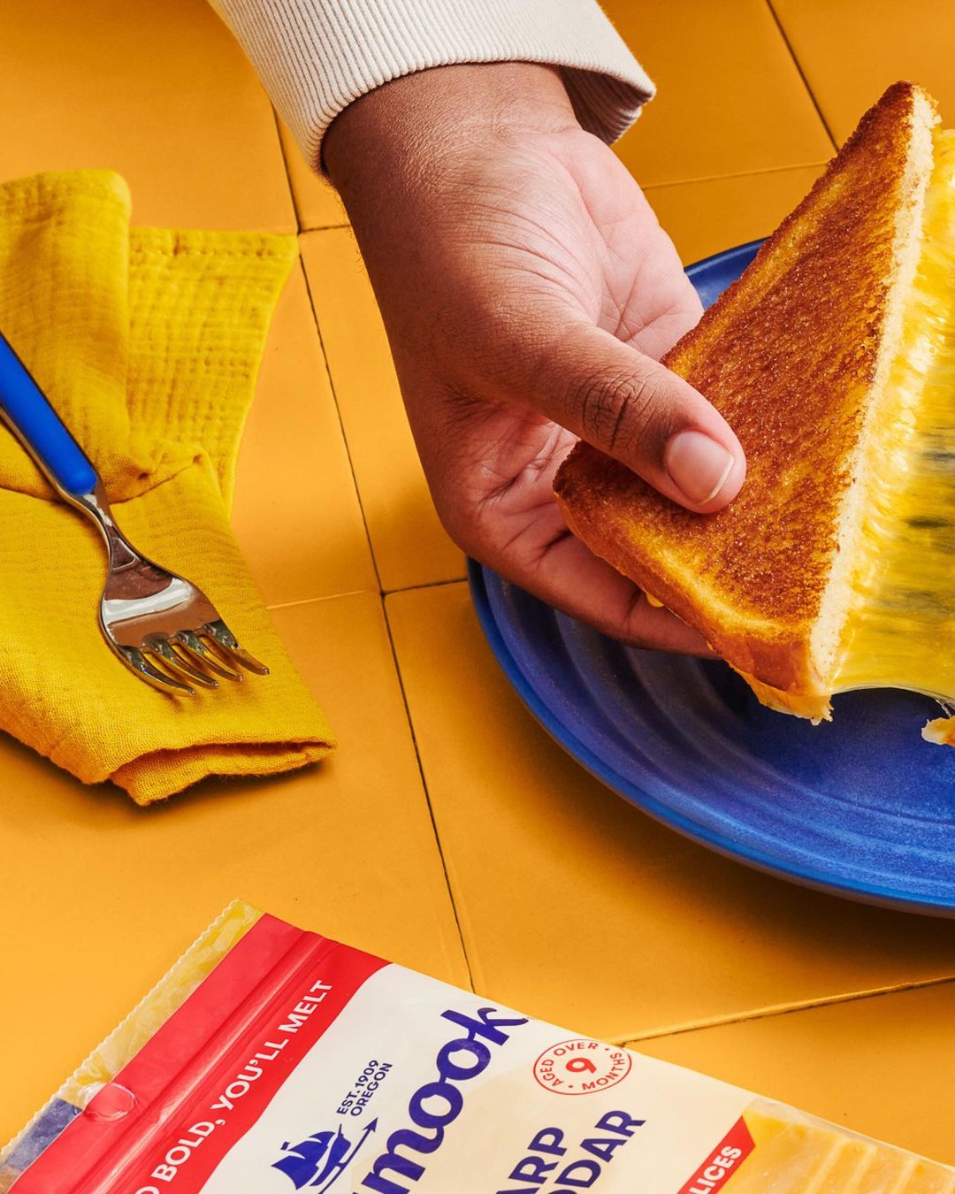 Close-up of a grilled cheese sandwich being held over a blue plate, with a fork, yellow towel, and package of cheese slices visible.
