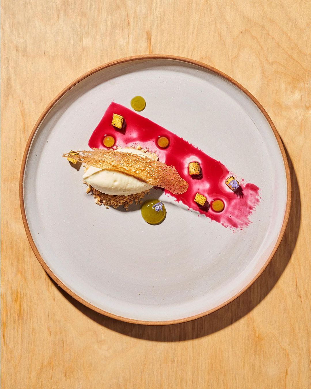 Overhead shot of artful dessert presentation on a round white plate with a beige rim, featuring white cream, red sauce, and edible garnish.