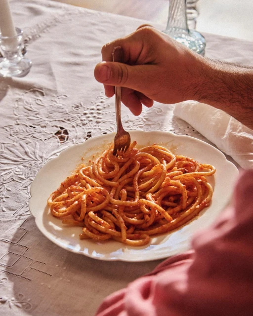 Overhead view of pasta with red sauce on a white plate being eaten with a fork by a person sitting at a table with white tablecloth.