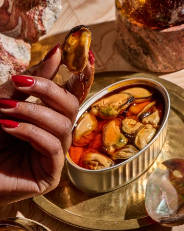 Hand holding canned mussels in oil on a golden tray, illuminated with warm lighting, creating a rich, textured food presentation.