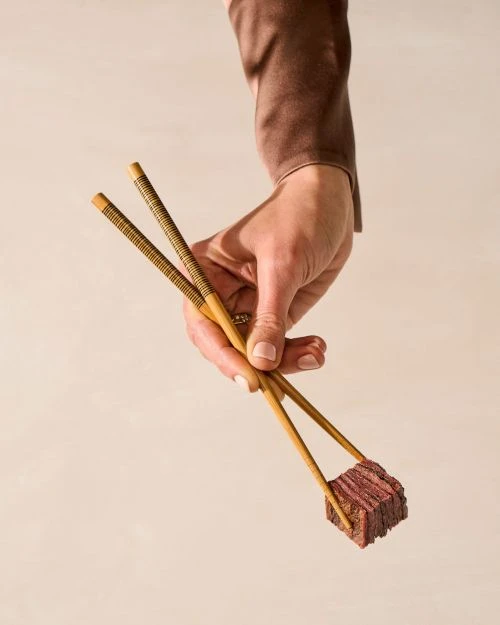 Close-up of a hand holding a cube of meat with chopsticks against a light background in a studio-lit setting, food photography.