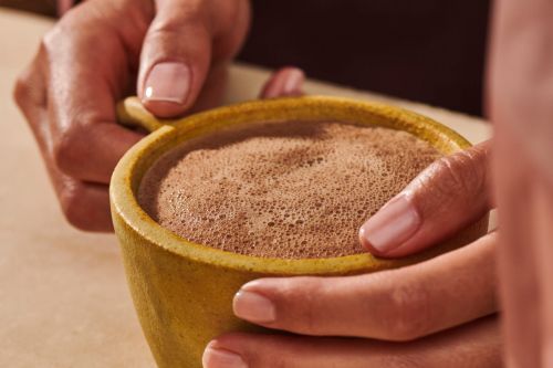Close-up of a person holding a yellow mug filled with frothy hot chocolate, set indoors.