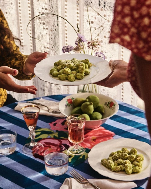 Two people passing plates of gnocchi at a table set with striped tablecloth, drinks, flowers, and a bowl of lemons in an indoor setting.