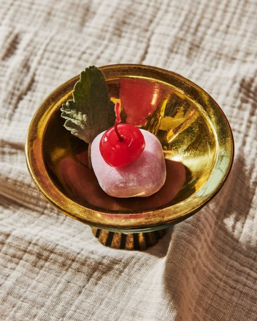 Close-up of a pink mochi dessert with a bright red cherry on top, presented in a shiny gold bowl set against a textured fabric.