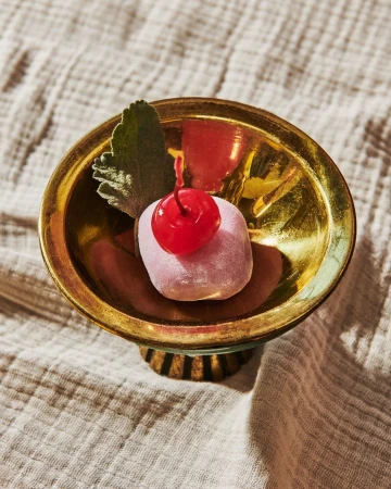 Close-up of a pink mochi dessert with a bright red cherry on top, presented in a shiny gold bowl set against a textured fabric.
