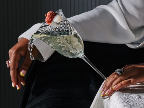 Elegant close-up of a cocktail glass with a lychee garnish, held by a woman wearing rings, against a dark grey background.