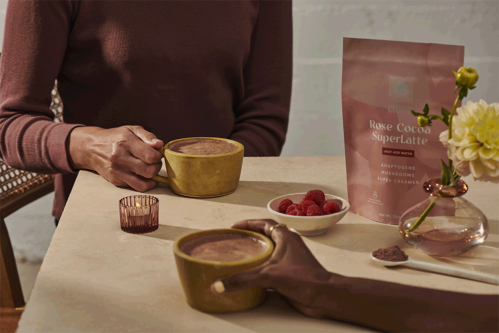 Close-up of two people at a table holding yellow cups of chocolate drink, with raspberries, flowers, and a drink mix bag.