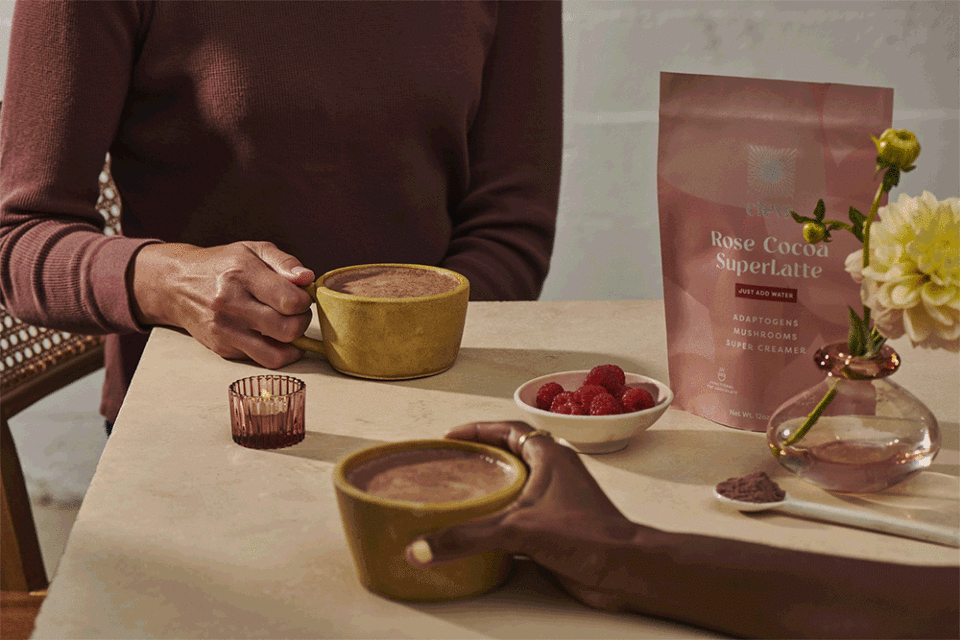 Close-up of two people at a table holding yellow cups of chocolate drink, with raspberries, flowers, and a drink mix bag.