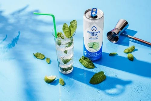 Can of Positive H2O sparkling cucumber water next to a cocktail glass filled with sparkling water, lime, and mint on blue surface.