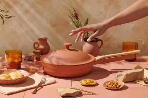 Still life with a terracotta cooking pan on a pink table surrounded by various dishes, glasses, and decorative objects in warm lighting.
