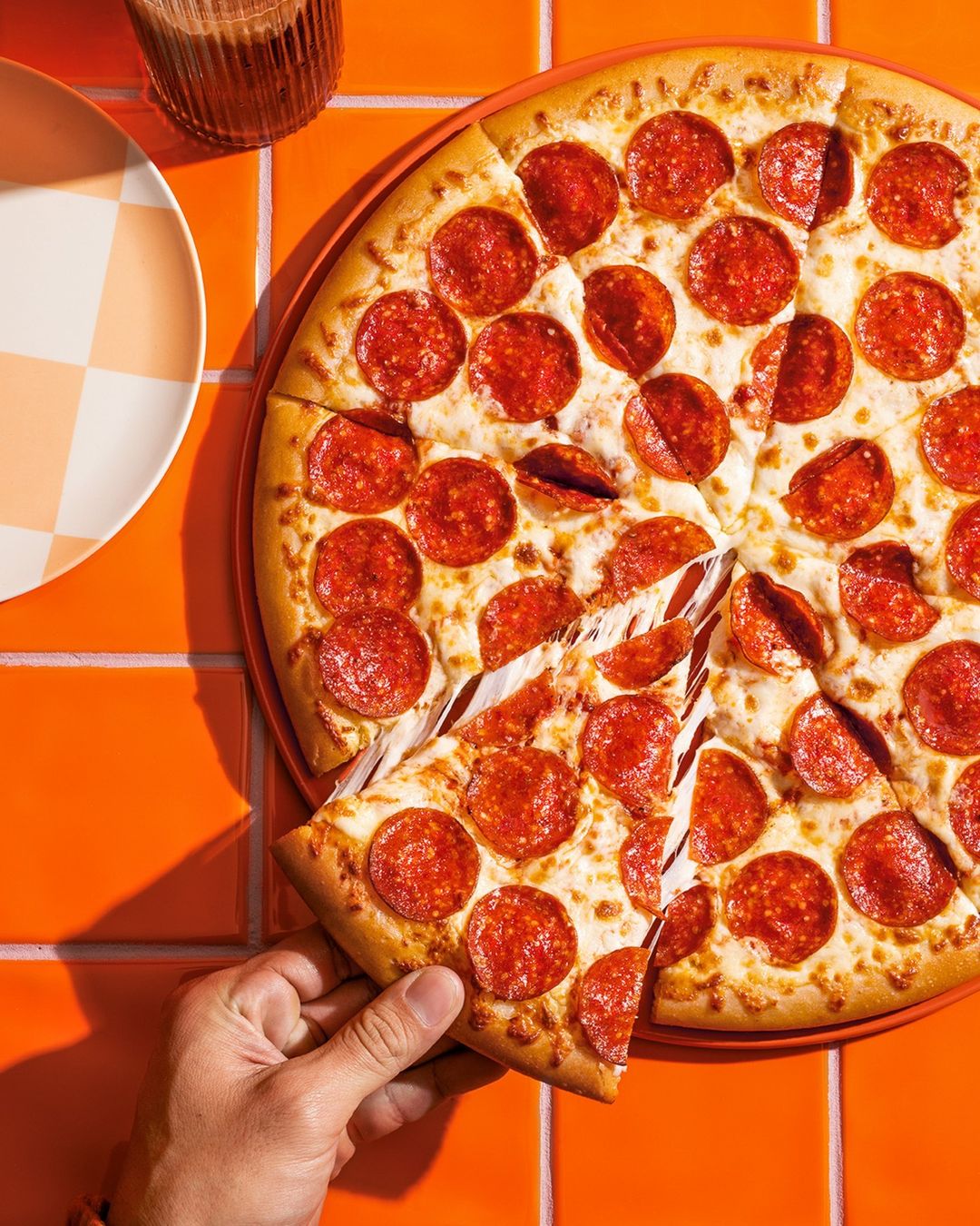 Overhead view of a pepperoni pizza on an orange tile surface, with a hand taking a slice. A plate and glass are visible.