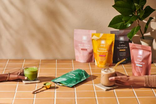 Hands hold latte drinks beside latte mix packets on a tiled surface with a potted plant and beige wall in the background.